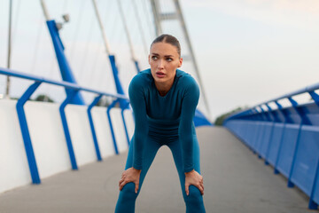 Young tired woman runner in sports outfit resting after running on the bridge in the city in the evening, taking break and breathing