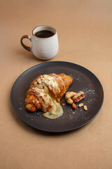 Croissant with filling peanuts and almonds with powdered sugar lies on black plate. Food photo cup brewed coffee on beige brown paper background, top view