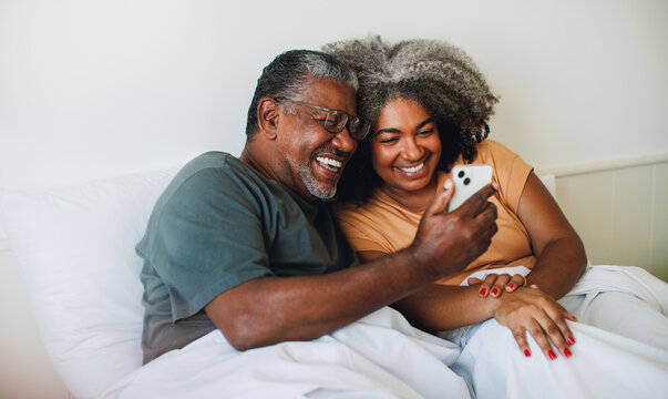 Connected Lifestyles: senior couple cuddling in bed while looking at phone together.