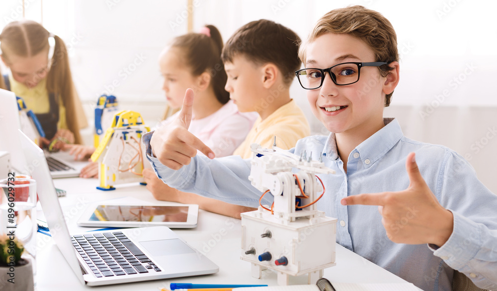 Aufkleber A young boy sits in a classroom setting with other students. He is smiling and pointing at a small robot he has built, demonstrating his learning and enthusiasm for STEM education.