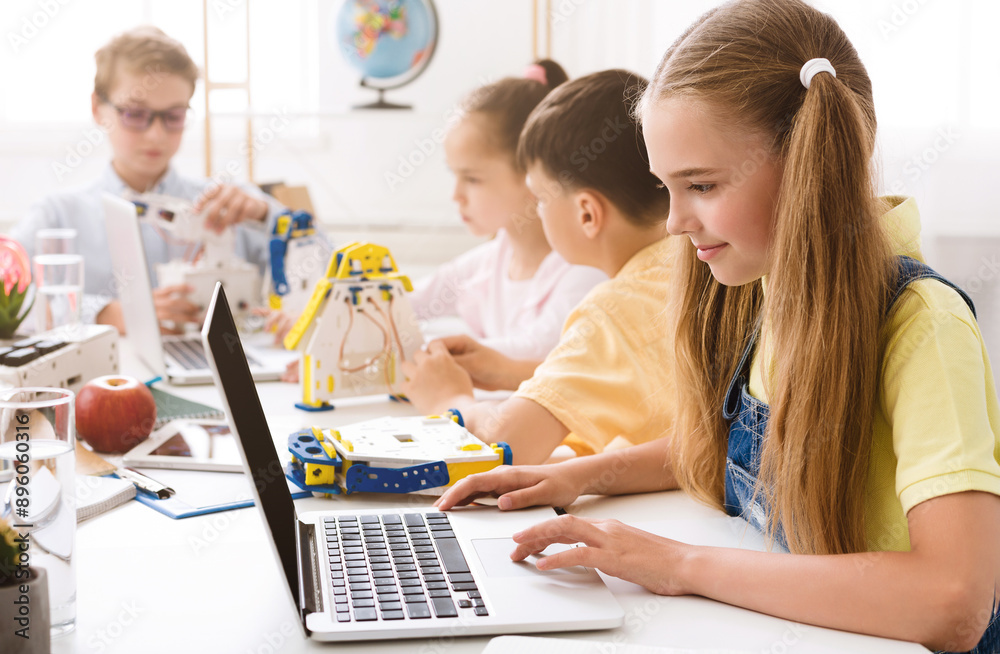 Poster a young girl sits at a table in a classroom, using a laptop. she is surrounded by other students who