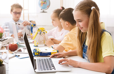 A young girl sits at a table in a classroom, using a laptop. She is surrounded by other students who are also working on projects. All kids focused on their work and appear to be enjoying themselves