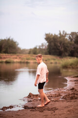 Kid boy teenager walking along the river bank and watches the water. Summer holiday concept.