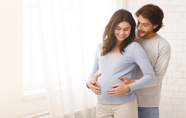 Fototapeta premium Loving man hugging his pregnant wife from behind standing near window at home, copy space