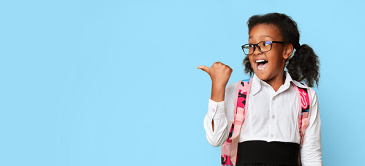Excited Black School Girl Pointing Thumb At Empty Space Standing Over Yellow Studio Background....