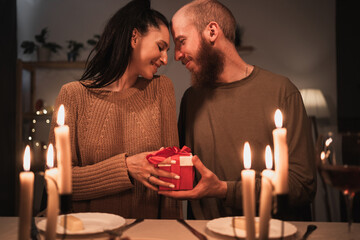 Happy young couple celebrating Valentines day having romantic dinner at home. Loving man giving gift box his woman.
