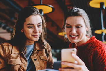 Positive female bloggers reading news in social networks using cellphone connected to wireless 4g internet, prosperous women looking at mobile screen searching information for learning indoors