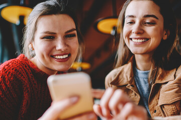 Two smiling female bloggers creating publication using photo application on mobile phone, happy...