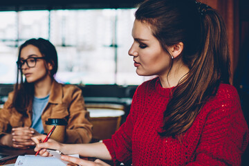 Young serious hipster girl searching information for homework and writing main theses in textbook using smartphone device sitting near female friend watching concentrated webinar via laptop computer