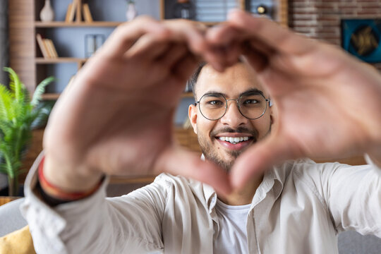 Smiling young man making heart shape with his hands during video call at home - Powered by Adobe