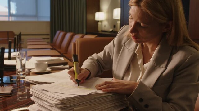 Mature business lady highlighting lines in paper on top of stack of documents with yellow textmarker while sitting at large wooden table in meeting room