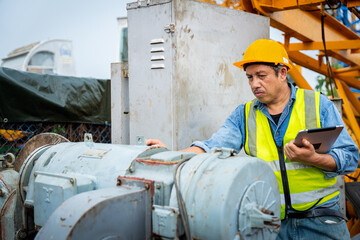 A man in a yellow safety vest is looking at a tablet while standing next to a machine. He is wearing a hard hat and he is focused on his work