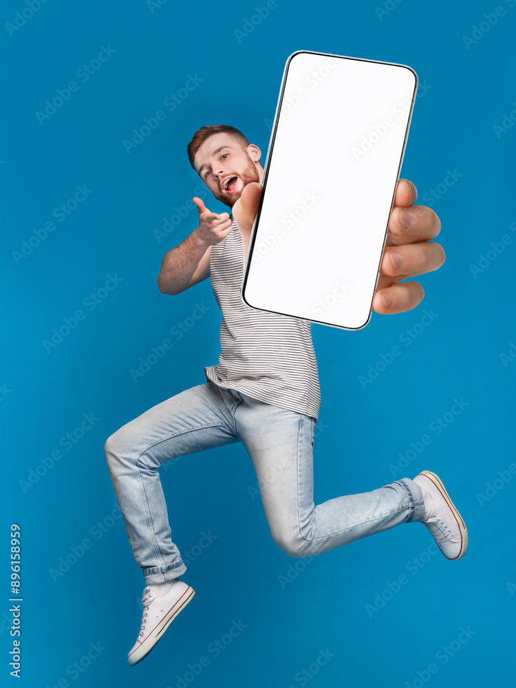 Poster emotional millennial redhaired guy pointing finger at camera, jumping with mobile phone on blue stud