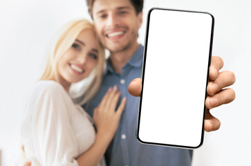 A couple is smiling and looking at the camera as a hand holds a smartphone with a blank white screen in the foreground.