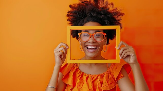 A delighted girl in an orange dress is holding an empty frame and gazing through it with a big grin, her eyes full of joy.