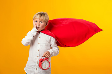 Surprised child girl plays superhero and holds alarm clock over yellow background. Education and success concept.