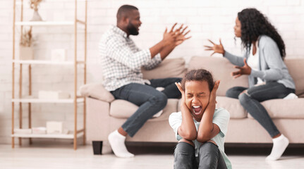 Defensive reaction. Upset little black girl sitting on floor, yelling and closing ears not to listen parents arguing, selective focus on child