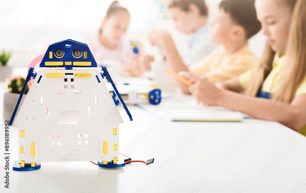 Aufkleber A close-up shot of a white and blue robot toy sitting on a table in front of a classroom of children. The children are out of focus in the background, and the robot toy is in sharp focus.