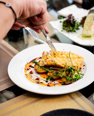 A close-up shot of a person cutting into a gourmet fish dish. A white plate holds a golden-brown fillet layered on top of roasted vegetables and a vibrant red sauce. The fish is garnished with fr