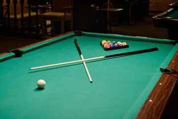 Wide and long table with green top and two crossed billiard cues lying between rack of balls and single white ball ready for game