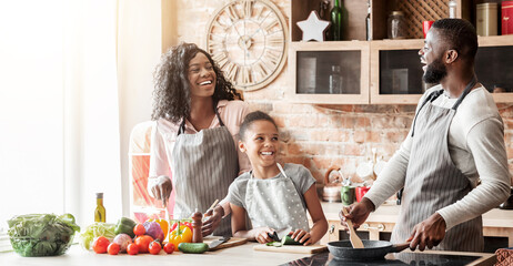 Happy black family of three laughing at kitchen while making healthy dinner, empty space