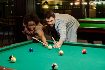 Young intercultural pool players with cues bending over wide billiards table with green top and looking at one of balls during game