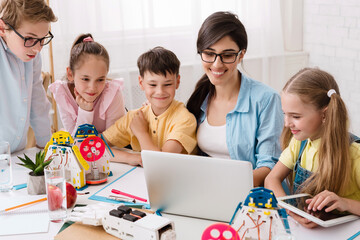 Elementary school students are working on a robotics project in their classroom. The students are gathered around a laptop and are looking at the screen intently. Their teacher is sitting beside them