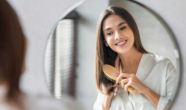 Beauty Routine. Pretty Woman Combing Her Beautiful Hair With Brush While Standing Near Mirror In Bathroom, Attractive Young Lady Looking To Her Reflection And Smiling, Selective Focus With Free Space