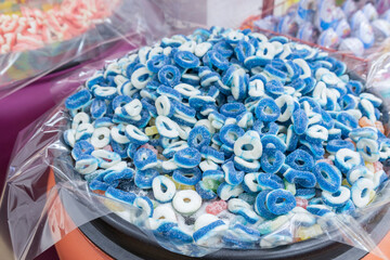 A close-up view of a container overflowing with blue and white gummy rings. The candy is displayed under clear plastic wrap, hinting at a candy store setting.