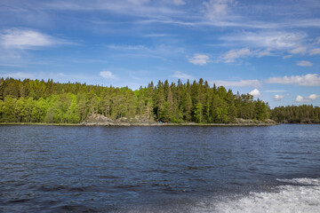 A view of a calm lake in Finland, with a lush, green forest lining the shoreline on a sunny summer day. The clear blue sky is dotted with white clouds.