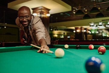 Young smiling man in casualwear and eyeglasses hitting white billiard ball with cue during game while bending over wide green table