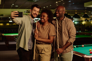 Young cheerful intercultural friends in casualwear looking at screen of mobile phone held by one of them during selfie