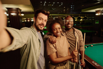 Happy young man with smartphone and his African American friends looking at you while standing in game room and enjoying leisure