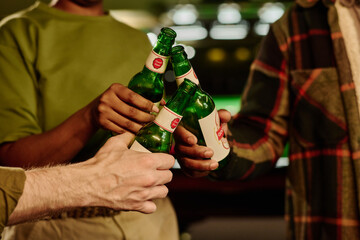 Hands of three young intercultural people clinking with bottles of beer standing in front of camera while toasting for Saint Patrick day