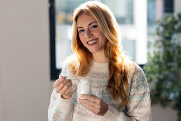 Happy beautiful woman eating yogurt while standing in living room at home