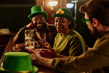 Young African American woman in green hat sitting by bar counter between two intercultural guys and talking to them