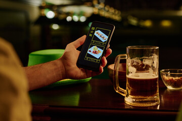 Smartphone with sushi photos on screen in hand of young modern man sitting by bar counter with mug of beer and choosing online food