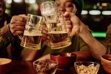 Hands of three unrecognizable young people toasting with beer in front of camera while enjoying celebration of Saint Patrick day in pub