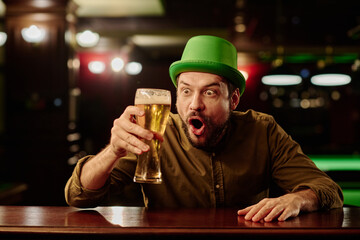 Young surprised man in green hat looking at glass of foaming beer while sitting by bar counter in Irish pub and celebrating Saint Patrick day