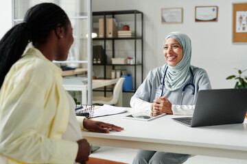 Obraz premium Young cheerful general practitioner in hijab looking at pregnant African American patient during medical consultation in office