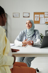 Young confident female general practitioner in hijab looking at pregnant patient sitting in front of her and giving her medical advice