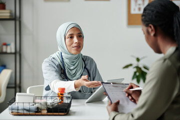 Young female doctor with tablet sitting in front of African American patient filling in and signing...