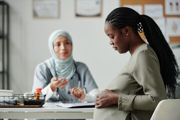 Profile view of young African American woman looking at her pregnant belly while sitting in front of her doctor in hijab in medical office