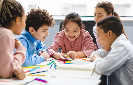 Technology And Pupils Concept. Group of excited multicultural happy junior children sitting at table and using smartphone, playing online mobile games. Modern Device, Gadget Addiction - Powered by Adobe