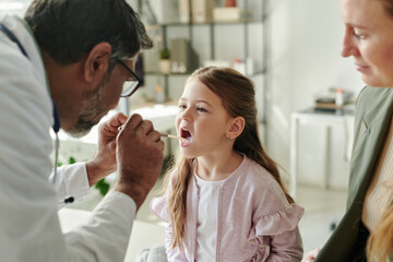 Obraz premium Cute little girl keeping her mouth open while mature pediatrician checking her throat with metallic medical spatula during consultation