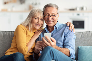 Happy Senior Couple Using Smartphone Browsing Internet Together Sitting On Couch At Home And Hugging. Cellphone Users Use Mobile Application. Older Spouses Texting On Phone. Communication Concept