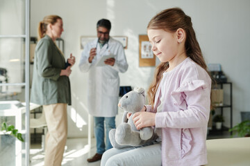 Fototapeta premium Side view of cute girl with grey soft toy sitting in front of camera in medical office and waiting for her mother consulting with clinician