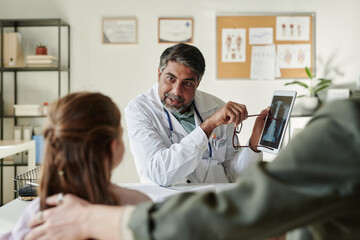 Confident mature pediatrician or radiologist pointing at x-ray image on tablet screen and looking at little patient during consultation