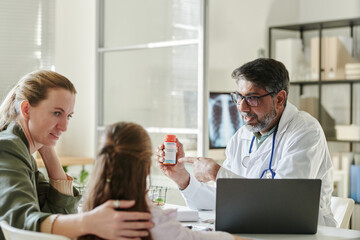 Confident male pediatrician pointing at bottle of pills or vitamins in his hand and looking at cute little patient visiting him with her mother