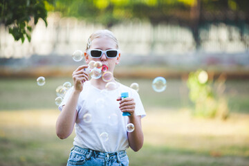 A little girl blows soap bubbles in the park in summer. Blonde girl in glasses, white T-shirt and...